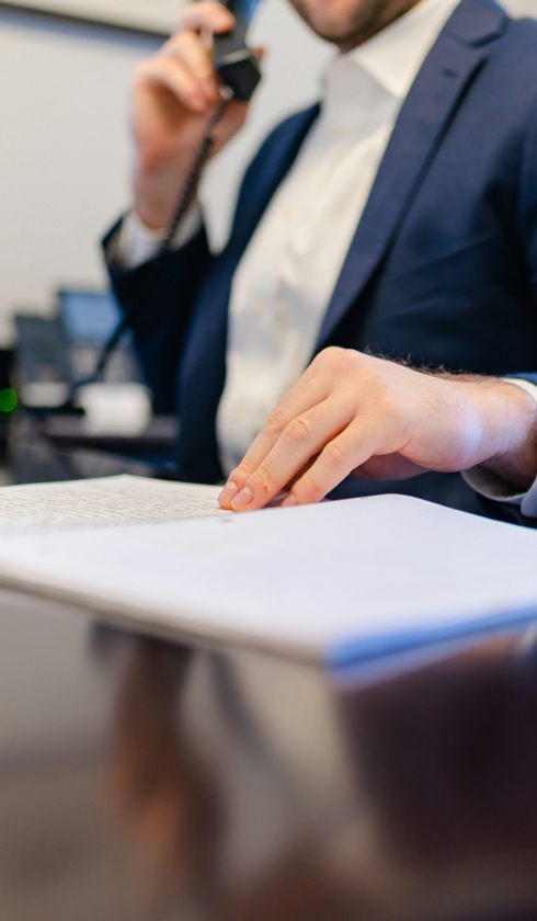 A person in a suit sits at a desk, holding a phone to their ear with one hand and reviewing damage calculations documents with the other. The image focuses on the person's hands and the paperwork.