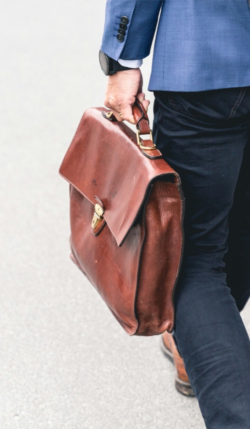 A person in a blue blazer and dark pants walks while holding a large brown leather briefcase by the handle, as if headed to review damage calculations, with a watch visible on their wrist. The background is a plain, light-colored surface.