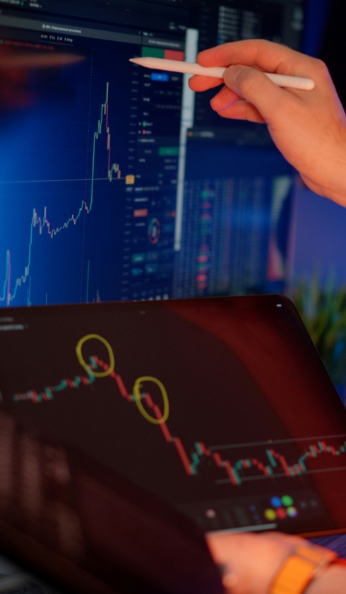 A person holding a stylus points at a computer screen displaying a financial chart, possibly analyzing damage calculations. Another device below shows a candlestick chart with two yellow circles highlighting specific data points.