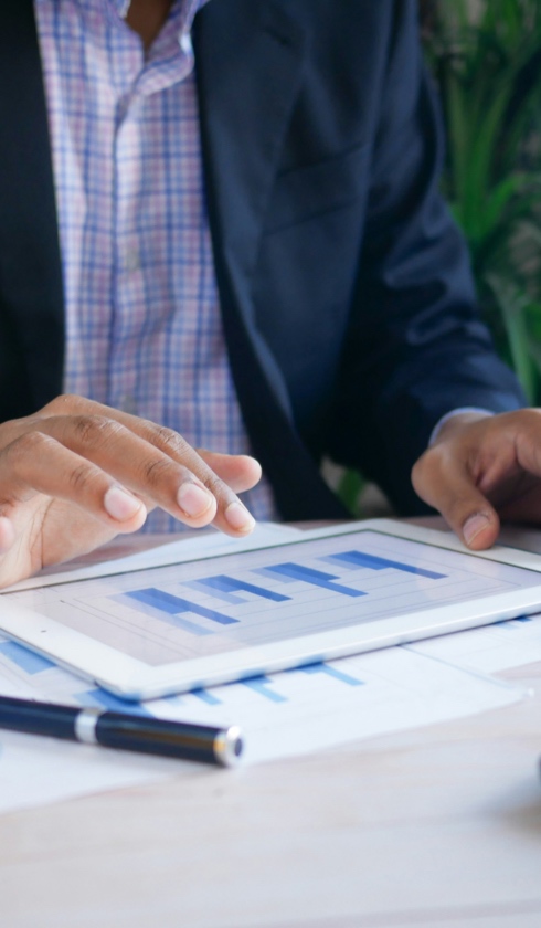 A person in a suit uses a tablet displaying bar graphs for damage calculations, with a pen and printed charts on the table.