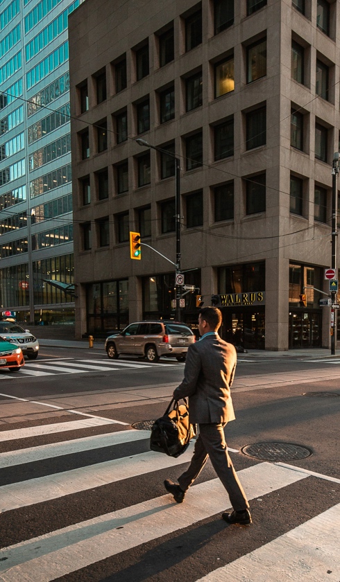 A man in a suit carrying a bag crosses a city street at a crosswalk, appearing deep in thought about damage calculations, with cars and tall office buildings in the background during daylight.