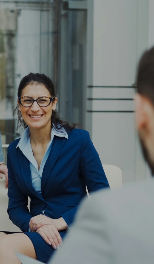 A woman in a blue suit and glasses is sitting and smiling during a conversation about damage calculations with another person in a modern office setting.