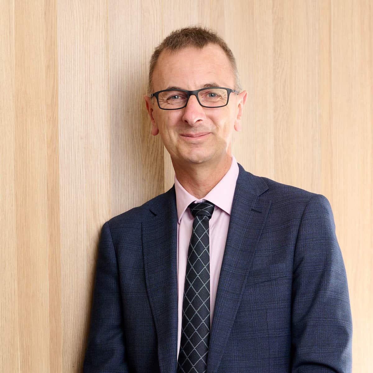 A man wearing glasses, a blue suit, a pink shirt, and a checkered tie is standing against a light wooden wall. He is smiling and has short hair.