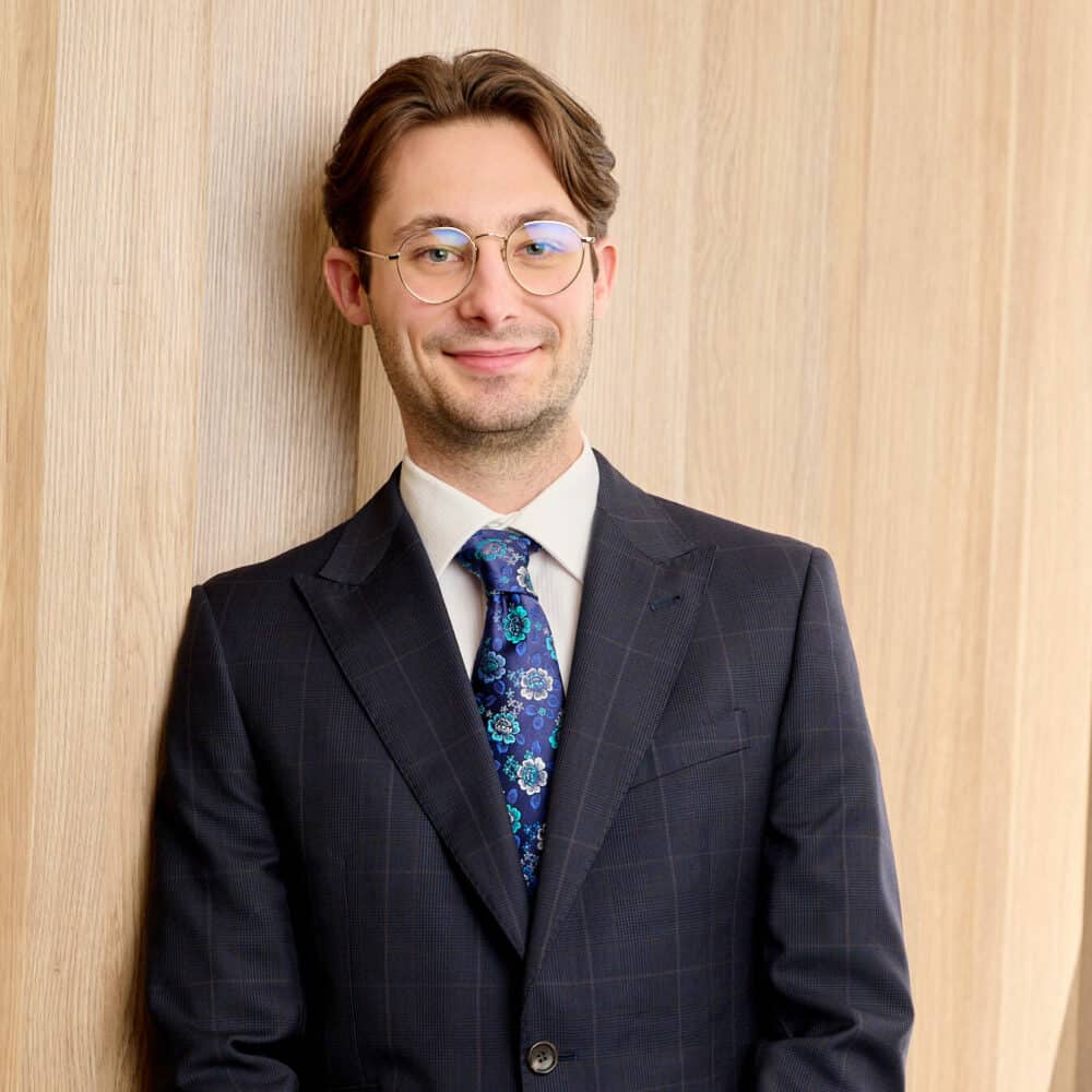 A person with glasses, wearing a dark checkered suit, white shirt, and a blue floral tie stands smiling against a wooden paneled wall, embodying the charisma of Zabard Hartmann.