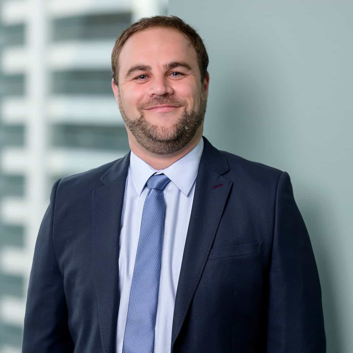 A man wearing a dark blue suit, light blue shirt, and blue tie smiles with his hands at his sides. He has short hair and a neatly trimmed beard. The background shows a blurred building with large windows.