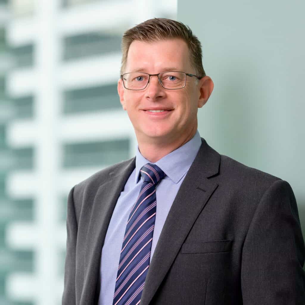 A man with short, light brown hair, wearing glasses, a dark suit, blue shirt, and striped tie is standing in front of a blurred, light-colored background. He is smiling and facing the camera.