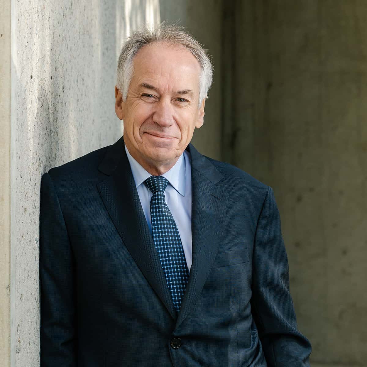 A man with gray hair, wearing a dark suit and a blue patterned tie, is smiling slightly while leaning against a concrete wall.