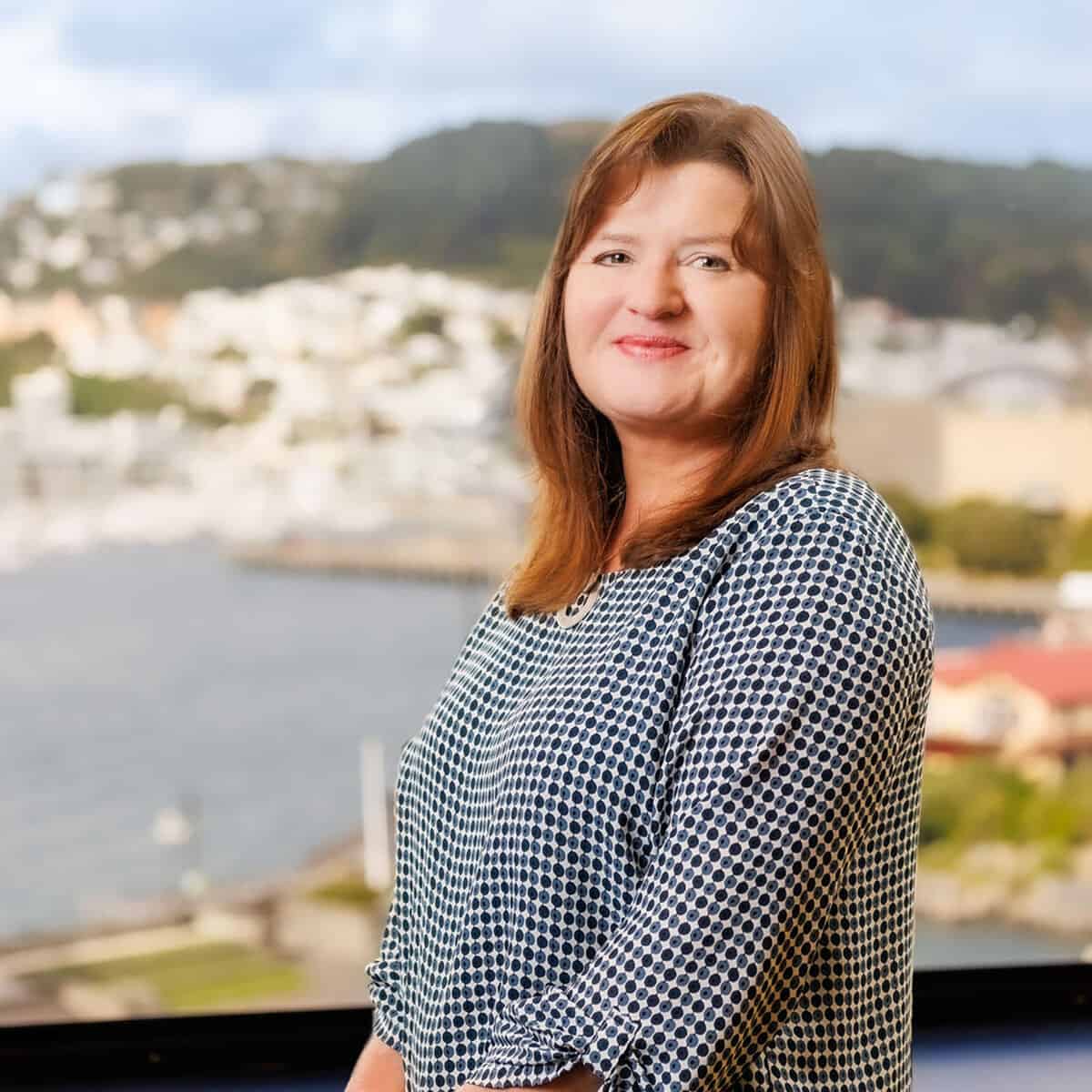 A woman with shoulder-length brown hair and a checkered black and white top stands smiling in front of a large window with a scenic view of hills, buildings, and water in the background.