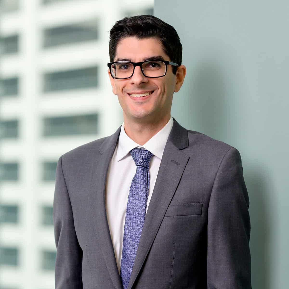 A man with dark hair and glasses is wearing a grey suit, white shirt, and blue tie. He is smiling and standing against a greenish-grey background. A tall, blurry building is visible in the background.