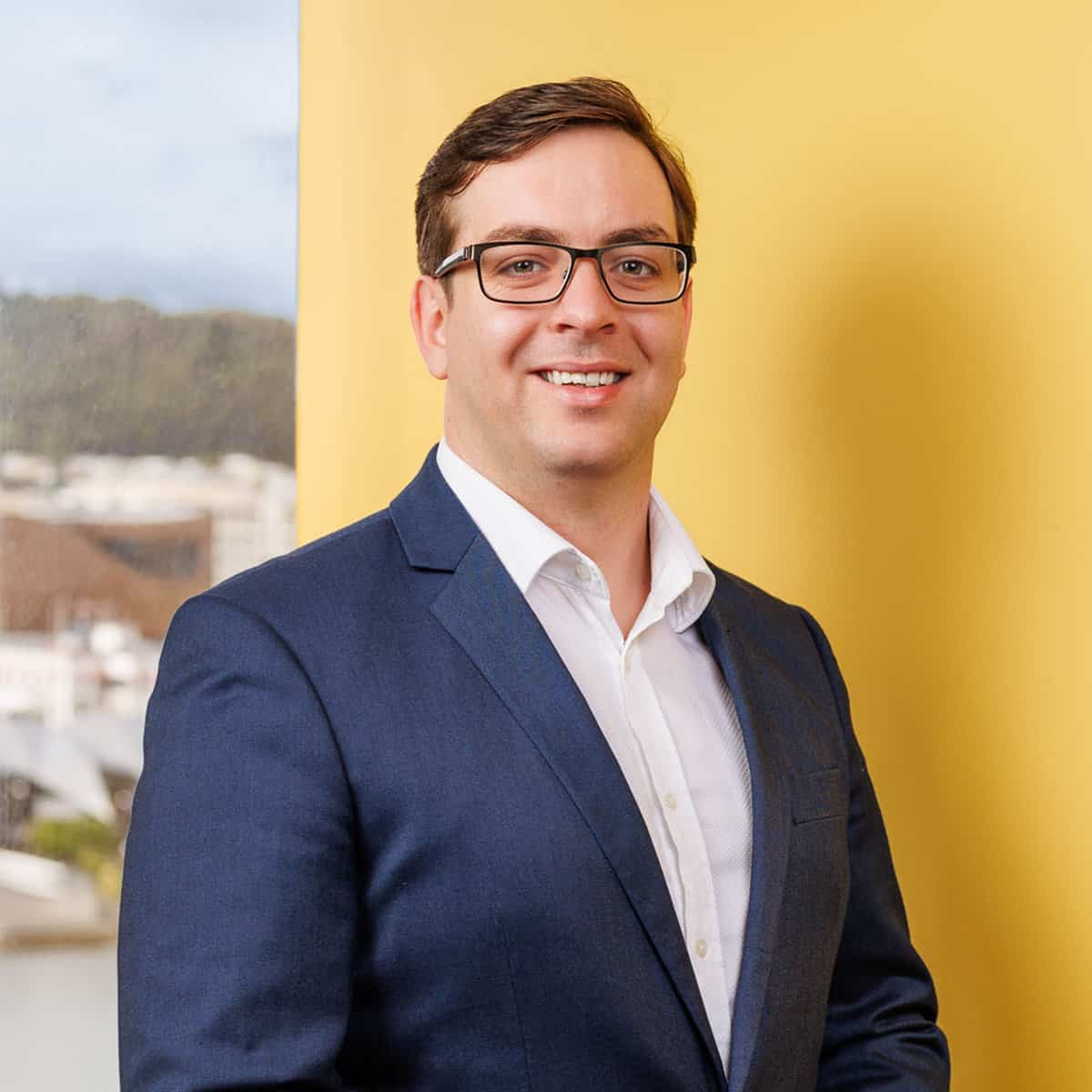 A man with short brown hair and glasses is smiling at the camera. He is wearing a navy blue suit and white dress shirt. The background features a yellow wall and part of a window showing an urban landscape.