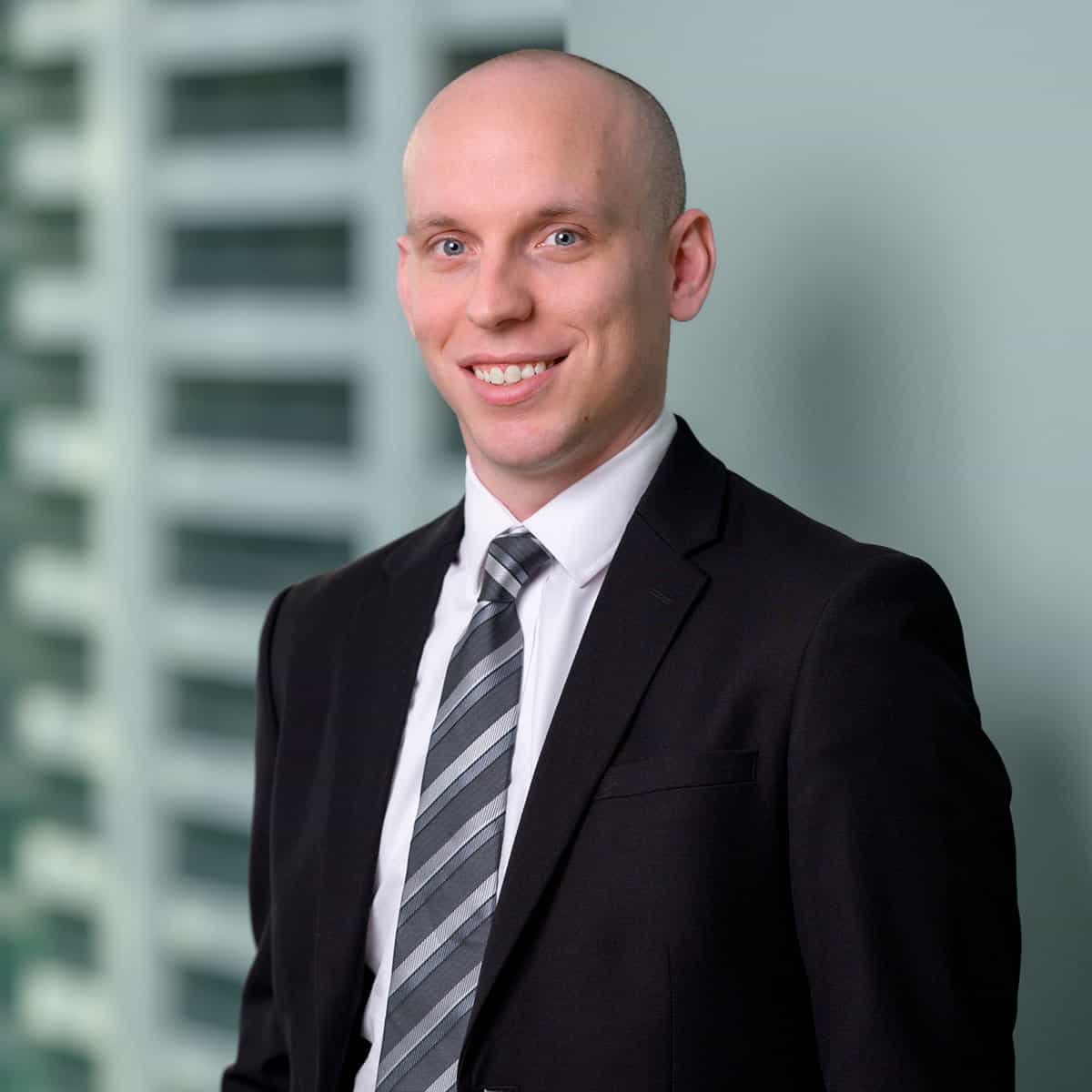 A smiling bald man wearing a black suit, white shirt, and gray striped tie stands in front of a blurred background with vertical lines.