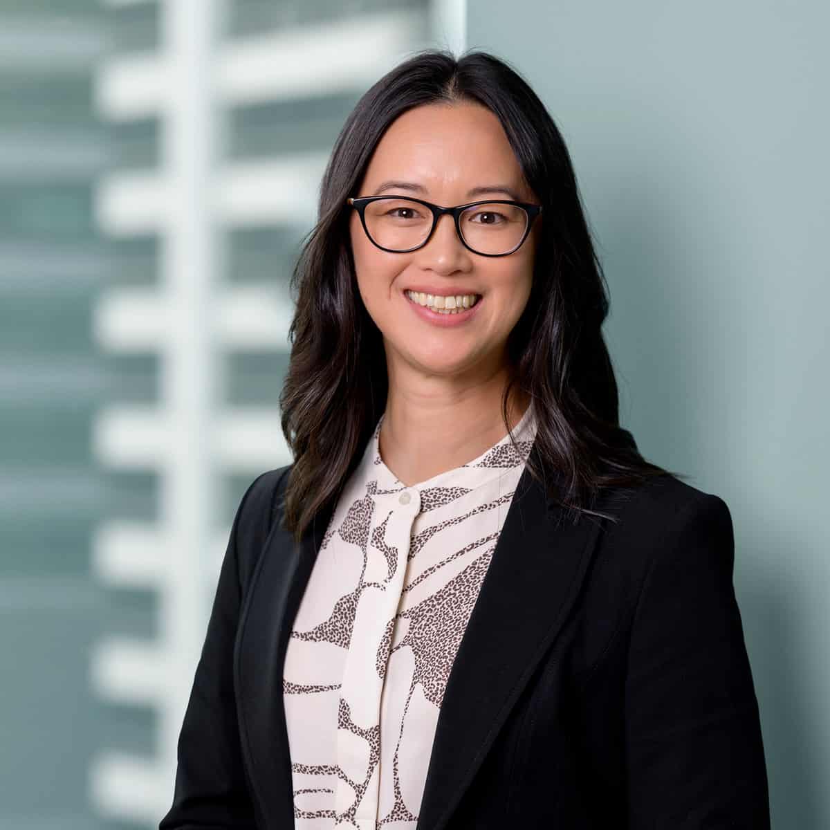 A smiling person with long, dark hair and glasses wearing a white patterned blouse and a black blazer. The background features a blurred geometric pattern with light gray and white tones.