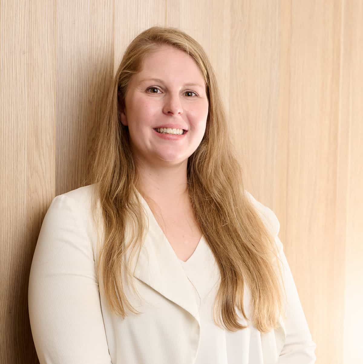 A person with long, light brown hair smiles while standing against a light wooden background. They are wearing a white or cream-colored top or blazer.