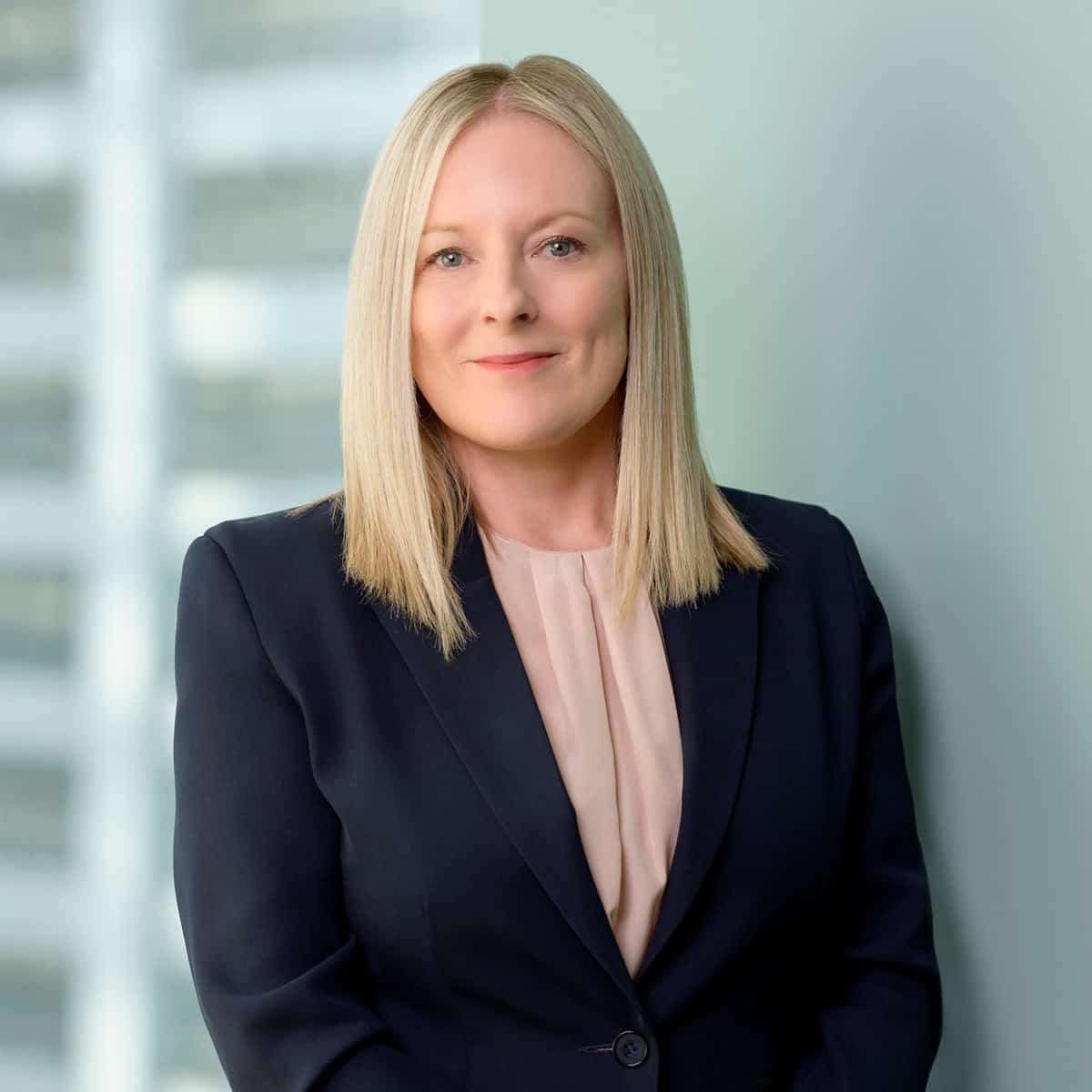 A woman with straight blonde hair, wearing a navy blazer over a light pink blouse, stands confidently in front of a blurred office building background. She has a slight smile and faces the camera directly.