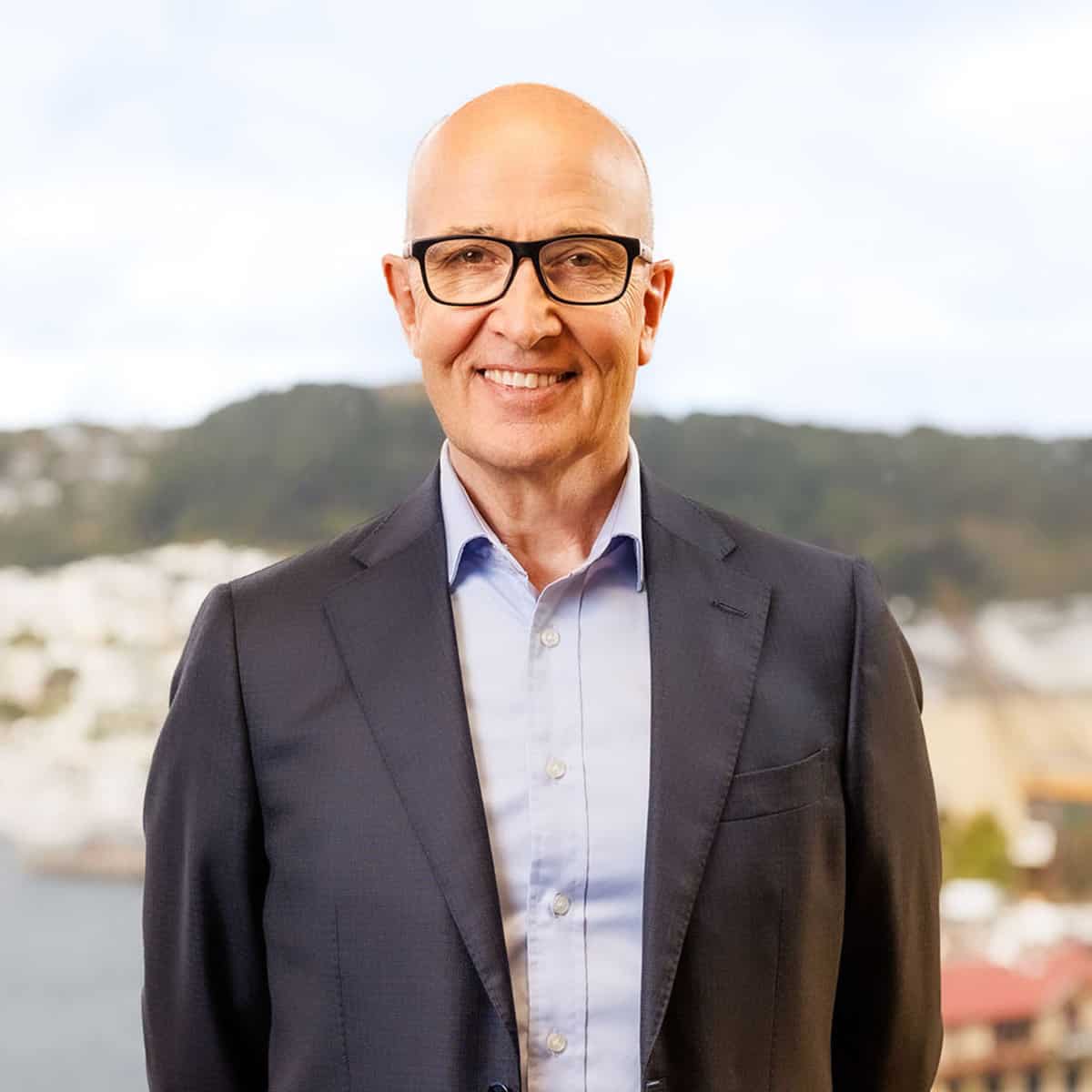 A smiling bald man wearing glasses, a blue dress shirt, and a dark blazer stands in front of a scenic background showcasing hills and a body of water. The sky is bright and overcast.