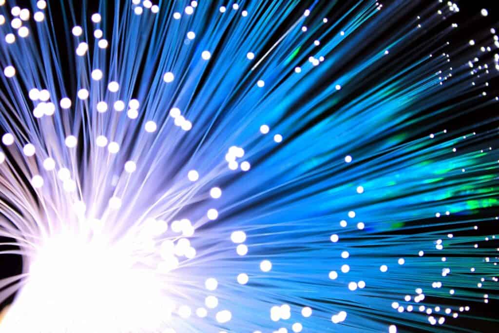 Close-up of glowing fiber optic cables. The image showcases numerous blue and green light strands fanning out with bright white light at the core, resembling a burst of energy. The glowing tips create a dynamic, futuristic visual effect.