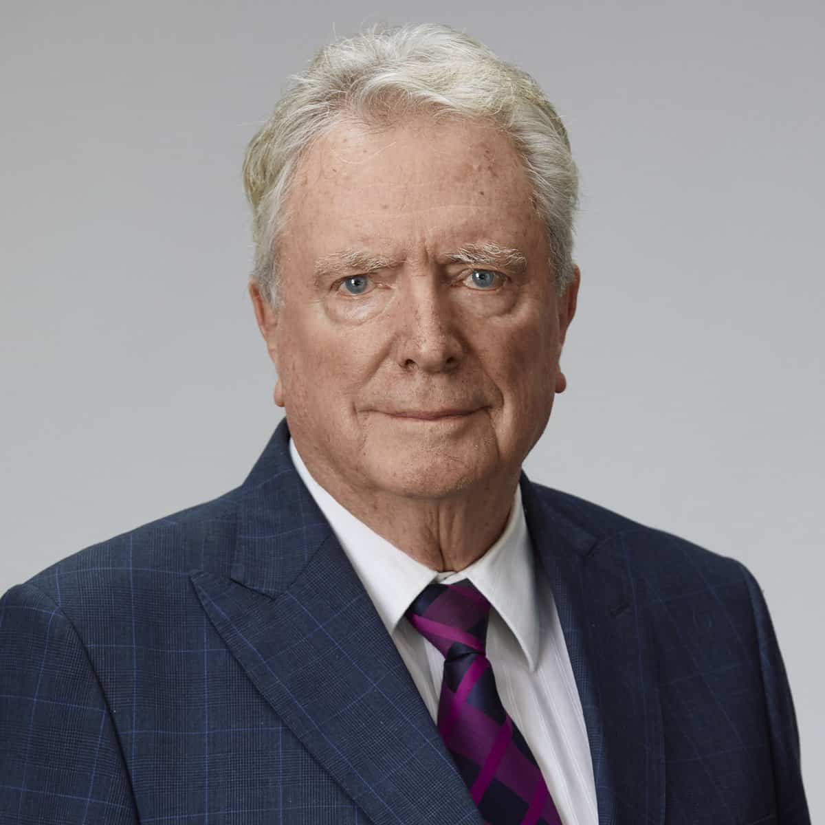 An older man with short gray hair is wearing a dark blue checkered suit, a white shirt, and a purple striped tie. He is standing against a plain background and looking directly at the camera with a neutral expression.