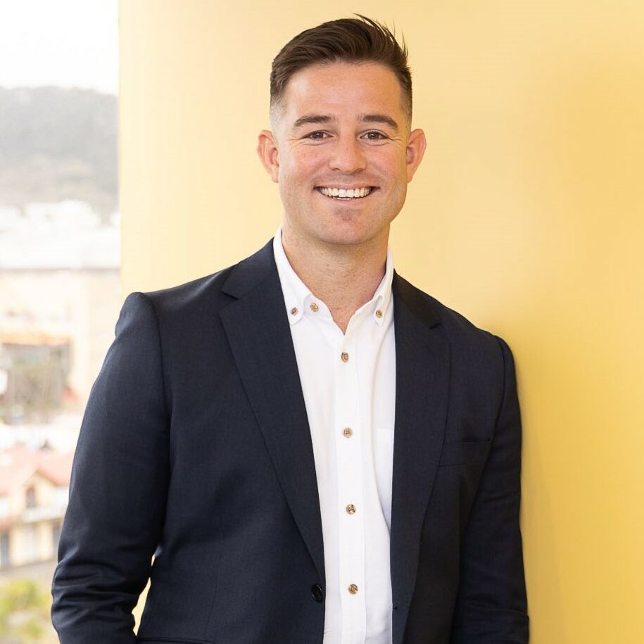 A smiling man with short dark hair stands against a yellow wall. He is wearing a dark blue blazer over a white shirt. His left arm is resting casually by his side. The background shows a blurred outdoor view.