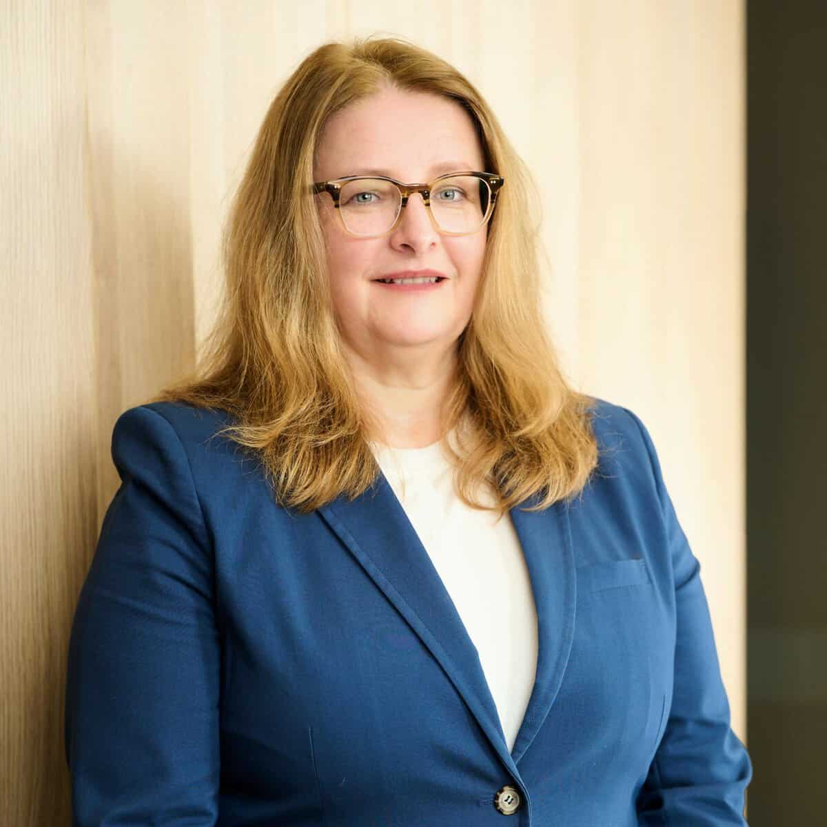 A woman with shoulder-length blonde hair and glasses is wearing a blue blazer over a white top. She is standing in front of a light-colored wooden wall and smiling slightly.