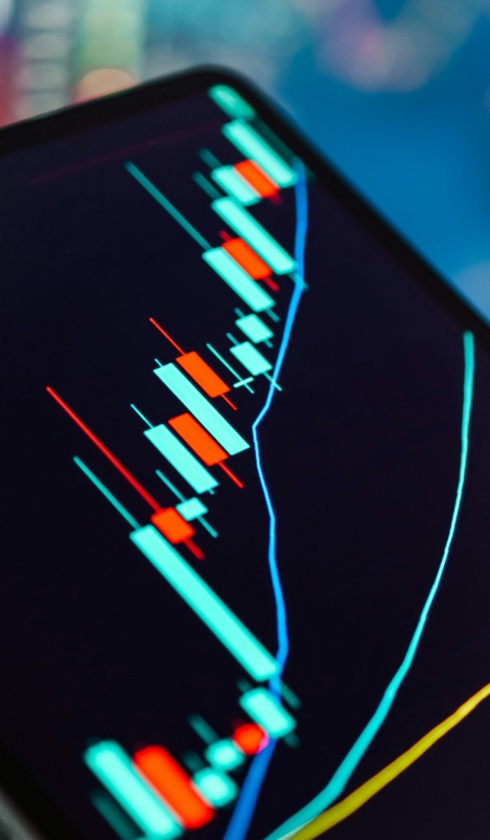 A close-up of a digital screen displaying a colorful candlestick chart with price and volume data, commonly used for financial or stock market analysis, on a dark background.