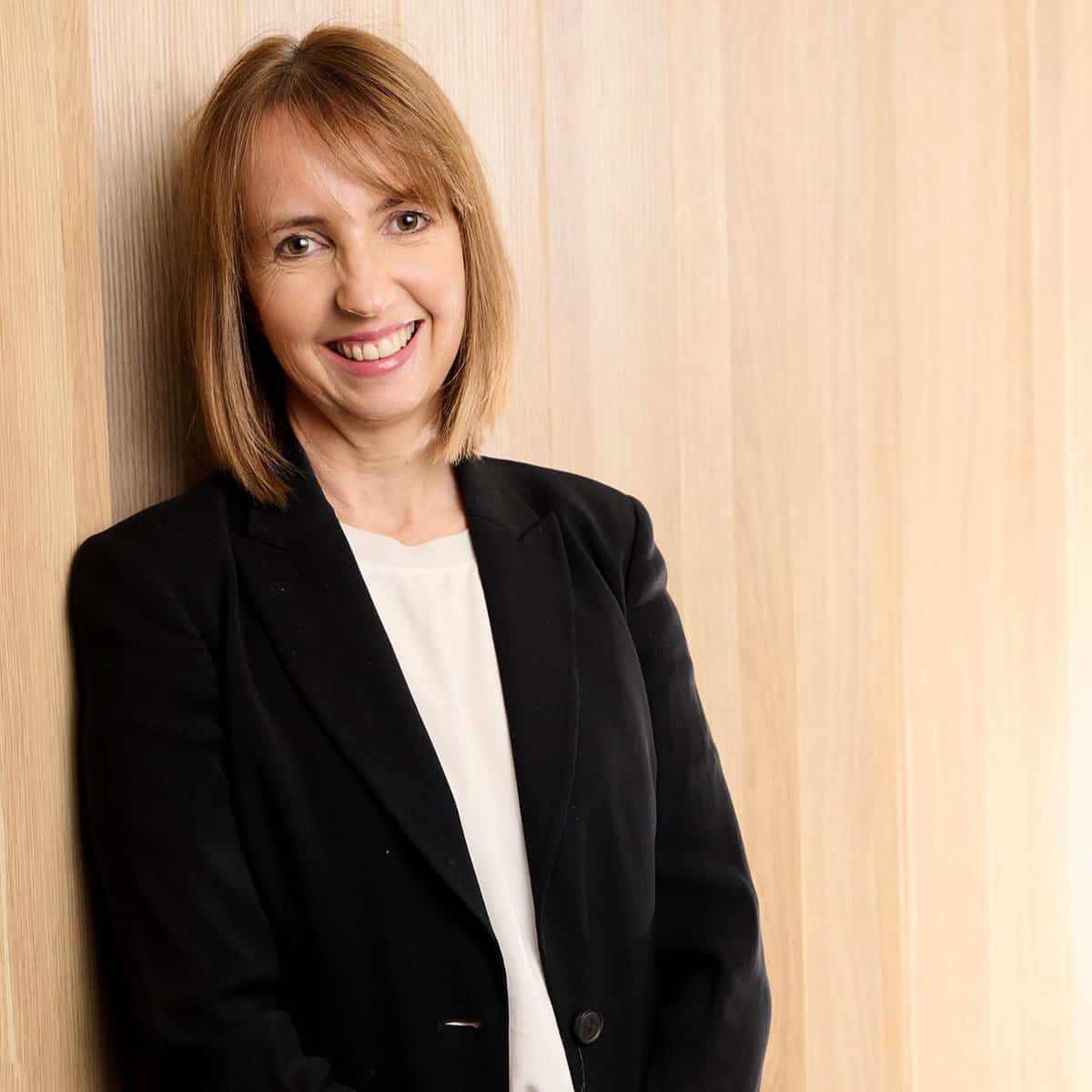 Sue, a friend with shoulder-length light brown hair, is smiling and leaning against a light wooden wall. She is wearing a black blazer over a white blouse.