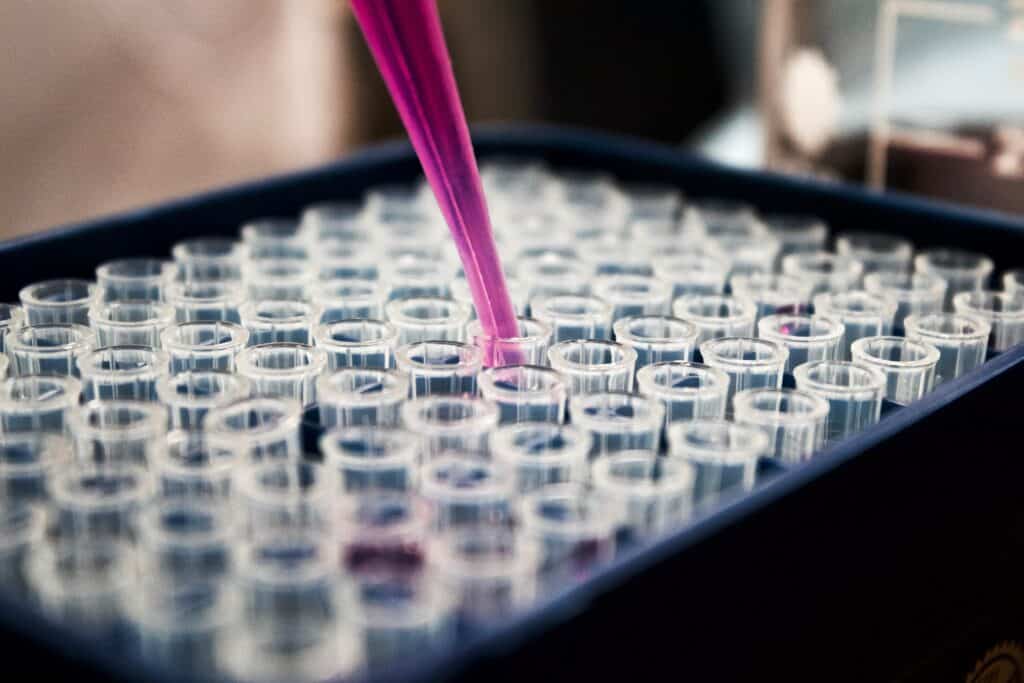 A close-up shows a pipette dispensing a pink liquid into a grid of small, transparent test tubes arranged in a tray, indicating a laboratory setting and possibly an experiment or analysis process in progress.