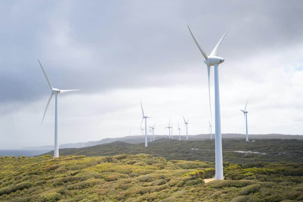 A series of white wind turbines stand amidst a green, hilly landscape under a cloudy sky. The turbines stretch into the distance, highlighting sustainable energy in a natural setting.