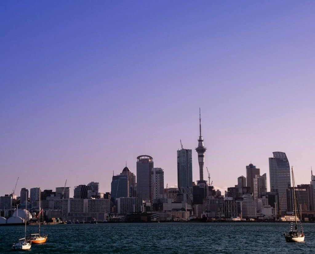 Panoramic view of a city skyline at dusk, with various skyscrapers and the prominent Sky Tower. The cityscape is reflected in the calm water in the foreground, where three sailboats are anchored. The sky is a clear gradient from blue to purple.