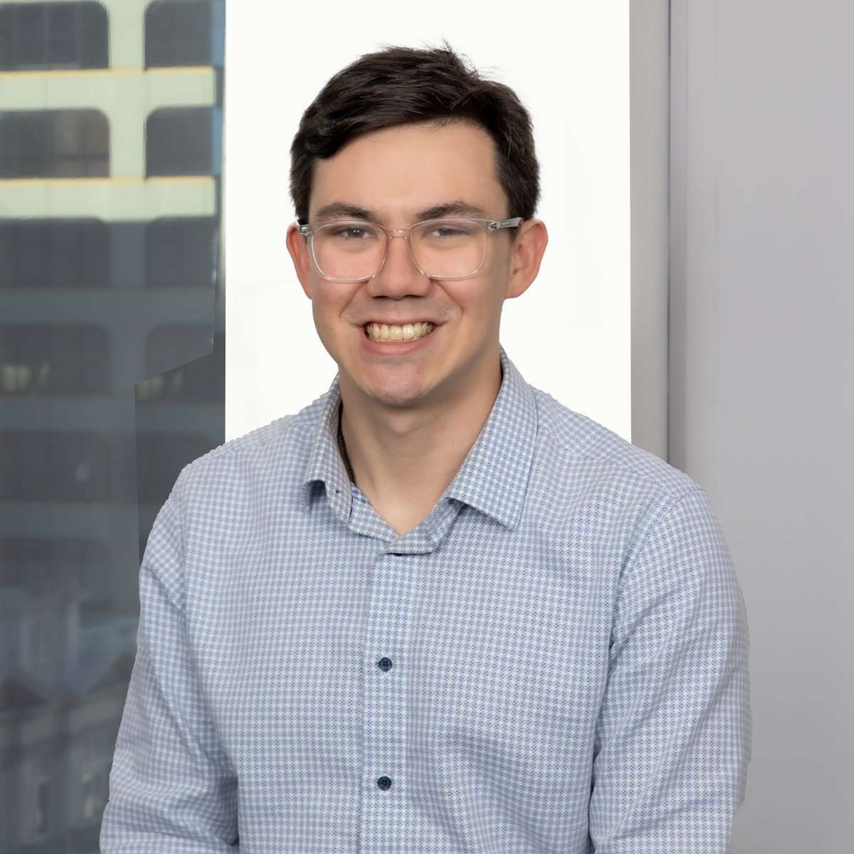 A person with short dark hair and glasses is smiling at the camera. They are wearing a light blue checkered shirt and are positioned in front of a window with buildings visible outside. The background includes a white wall section and part of a grey wall.