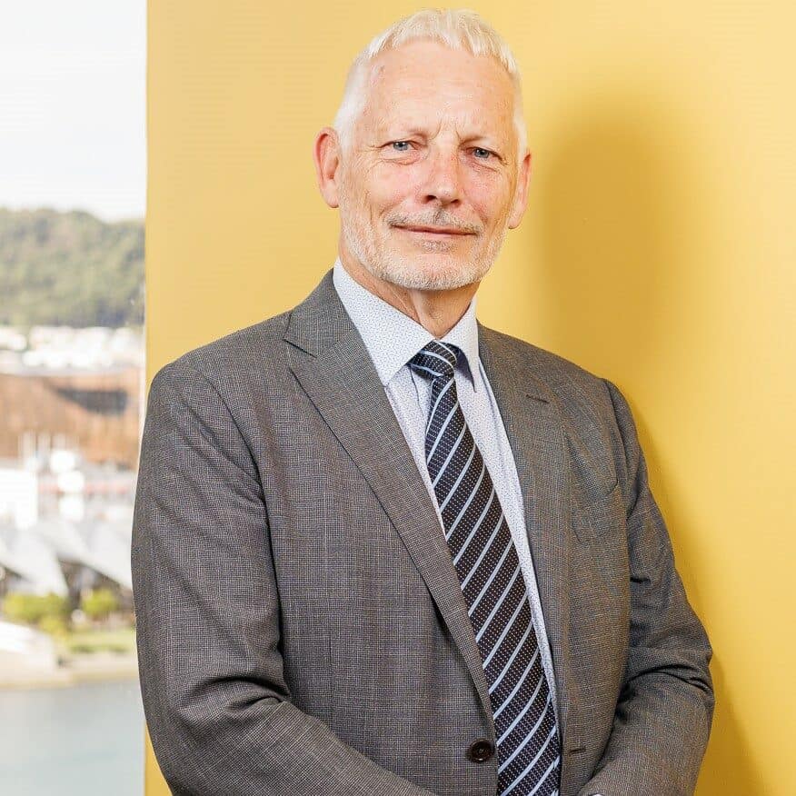 A man with short gray hair and a beard stands in front of a yellow background, wearing a gray suit, blue shirt, and striped tie. He has a neutral expression and his hands are clasped in front of him. The background also shows a blurry outdoor scene with trees.