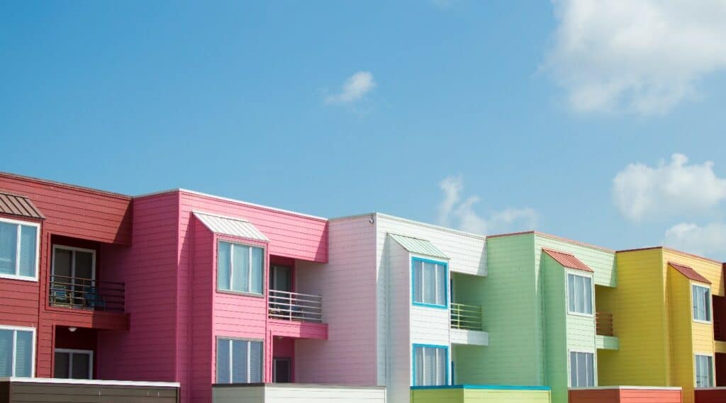 A row of colorful townhouses stands under a bright blue sky, each adorned in shades of pink, white, blue, green, and yellow. With large windows, small balconies, and matching awnings funded through housing agencies' support for social housing initiatives. Fluffy white clouds are scattered in the sky.