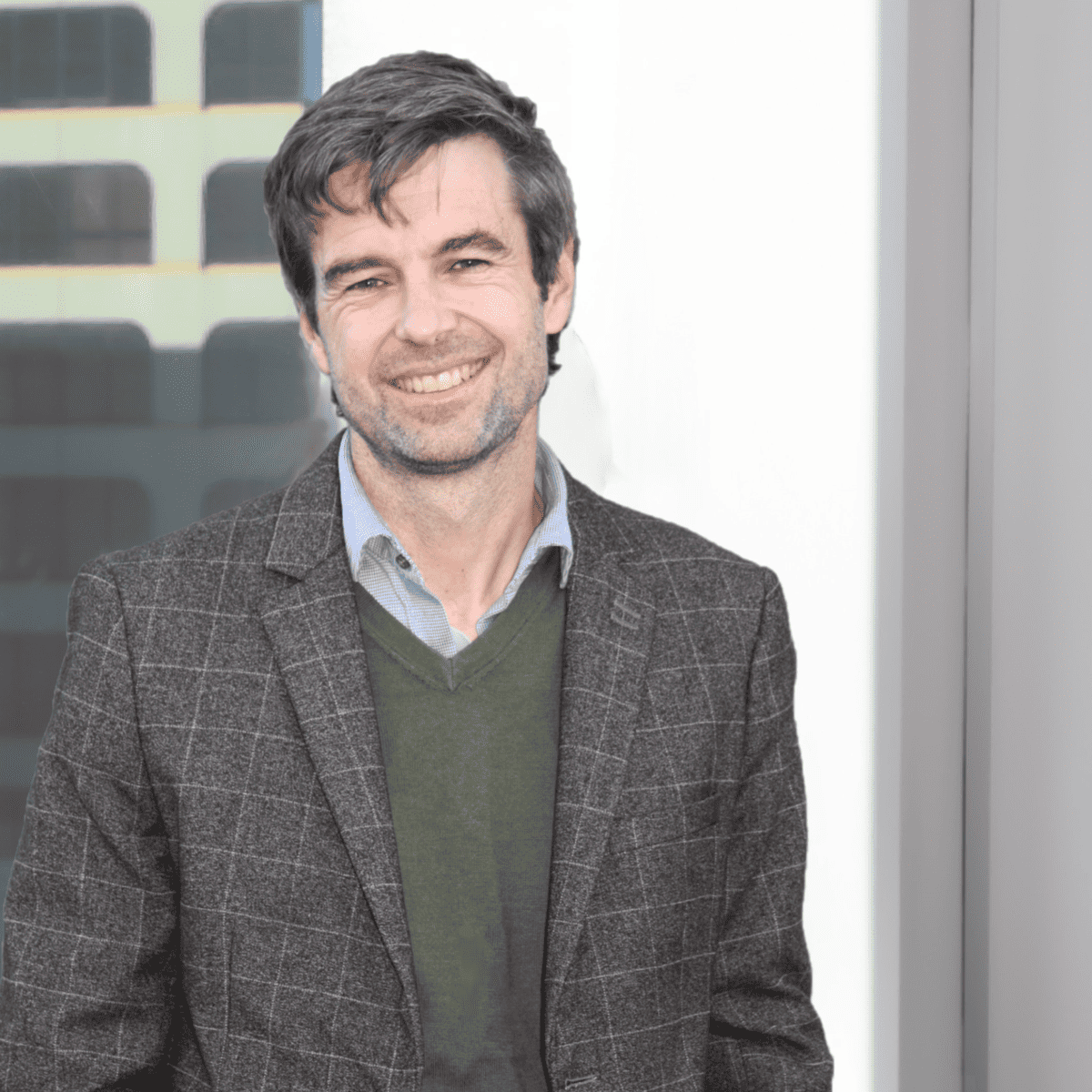 A man with graying dark hair and a short beard is smiling while standing in front of a window. He is wearing a gray checkered blazer over a green sweater and a light blue collared shirt. The background shows part of a modern building facade.