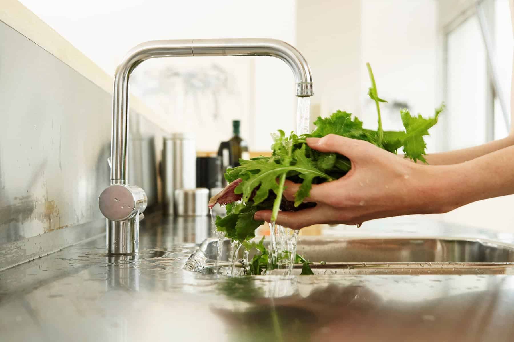 A person washing fresh green leafy vegetables under a running faucet in a modern kitchen. The water is splashing onto the stainless steel sink, and sunlight is streaming through a window in the background.