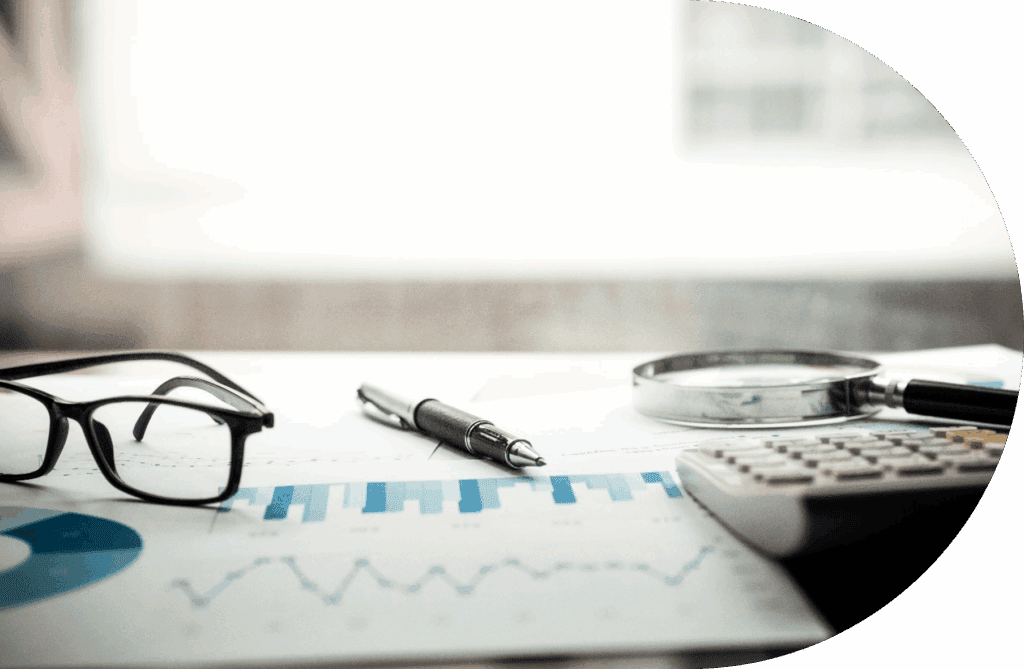 A close-up of a desk with eyeglasses, a pen, a magnifying glass, and a calculator on top of papers showing blue charts and graphs, suggesting financial management or data analysis.