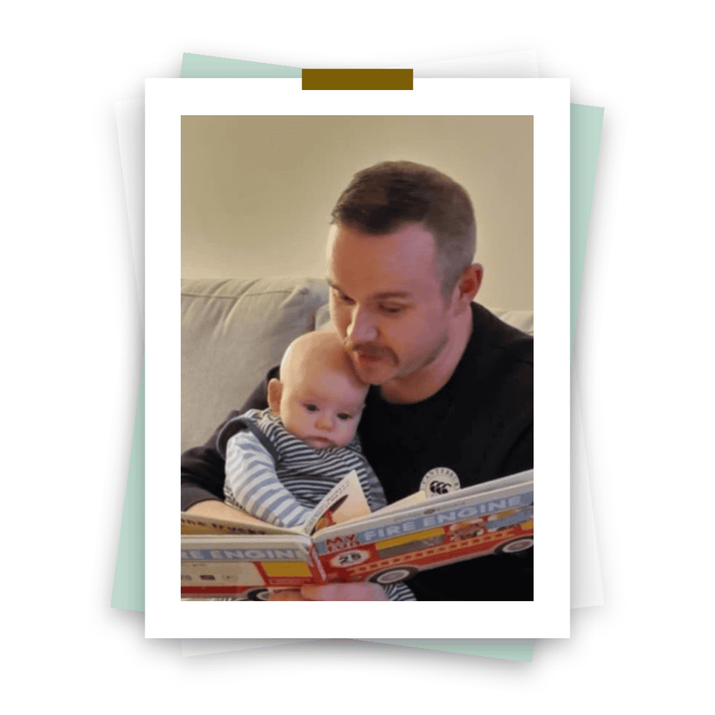 A man with short hair and a mustache is sitting on a couch, holding a baby and reading a book titled "My First Engine." The baby is nestled against the man, looking at the book. Both appear to be engaged in the story.