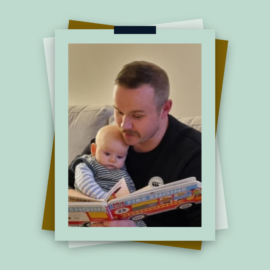 Dr Jamie O’Hare sits on a sofa reading a colourful book titled “My Fire Engine” to a baby on his lap. Both are intently focused on the book, creating a heartwarming staff spotlight moment that highlights Dr O’Hare’s dedication to nurturing young minds.
