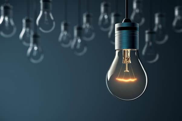 A close-up of a glowing light bulb hanging from the ceiling with several blurred non-lit bulbs in the background. The lighting creates a warm and focused effect against a dark blue backdrop.