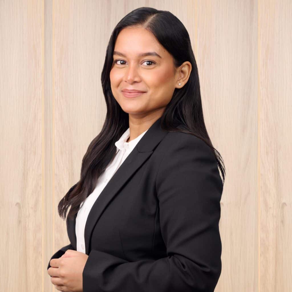 A person with long dark hair wearing a black blazer and white shirt stands smiling in front of a wooden panel background.
