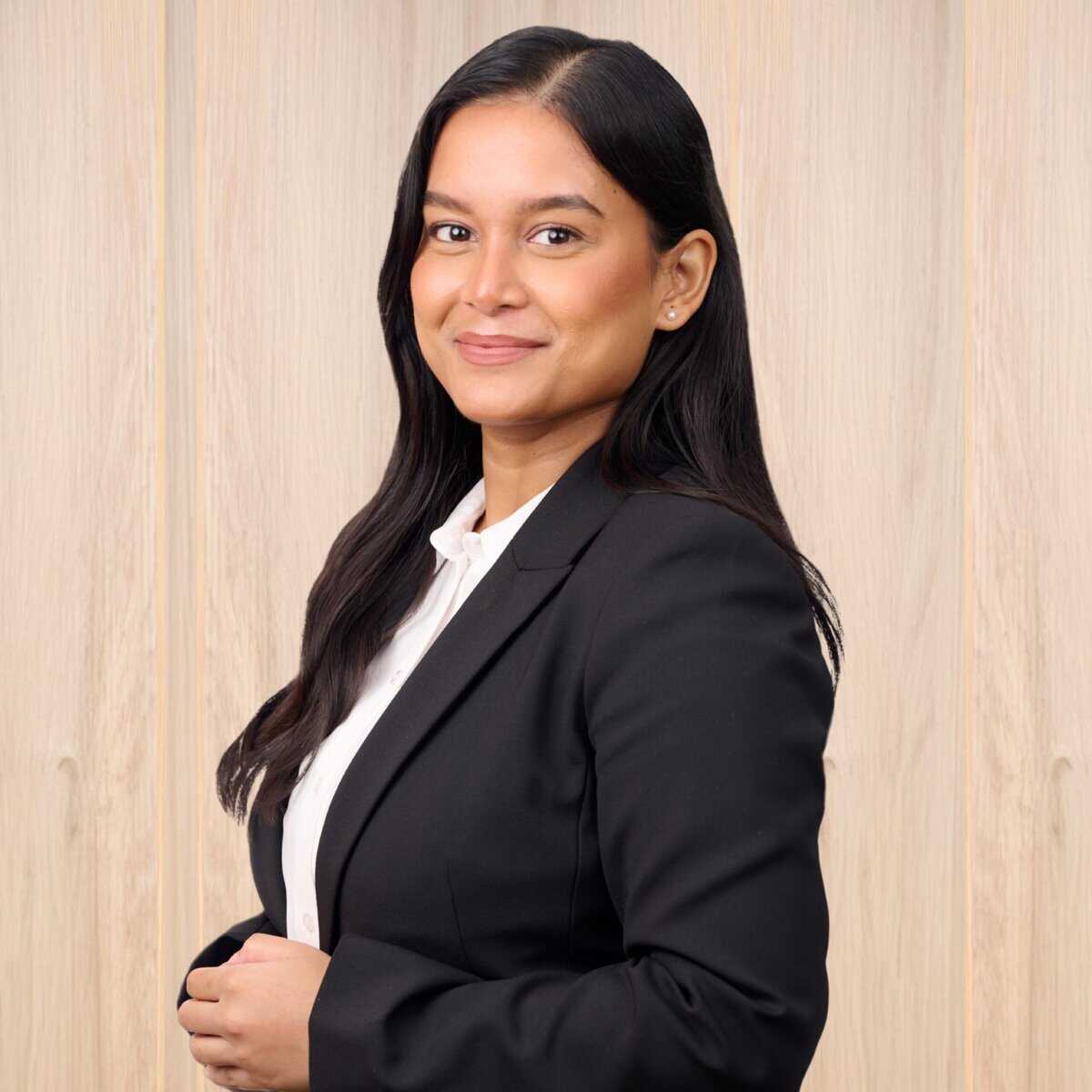 A woman with long dark hair, wearing a black blazer and white shirt, standing in front of a light wood background. She is smiling and looking towards the camera.