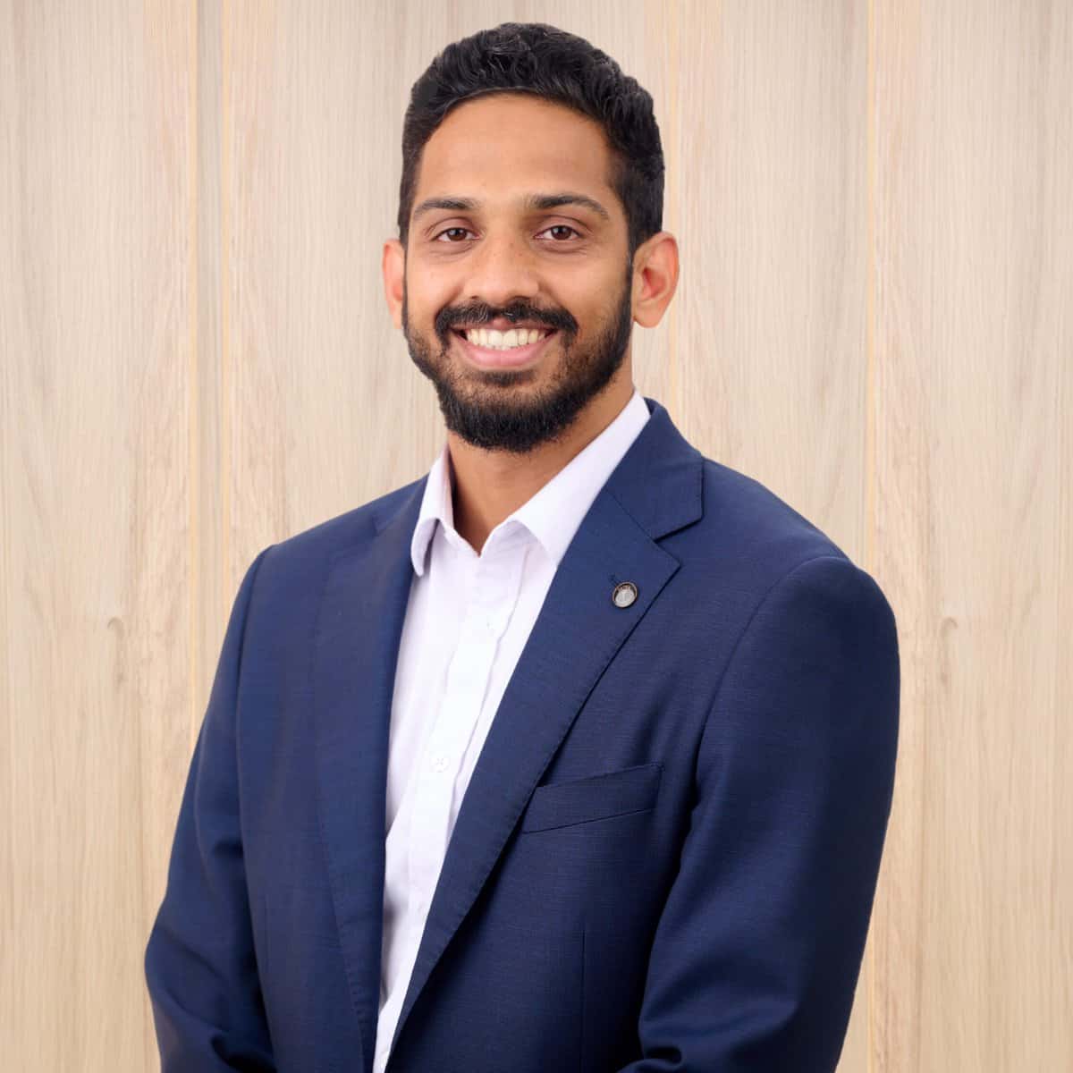 A smiling man with a beard is wearing a blue suit and white shirt. He is standing against a light wooden background.