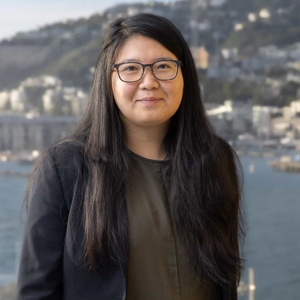 A woman with long black hair and glasses stands outdoors, wearing a dark blazer and olive shirt, with a waterfront city and hills in the background.
