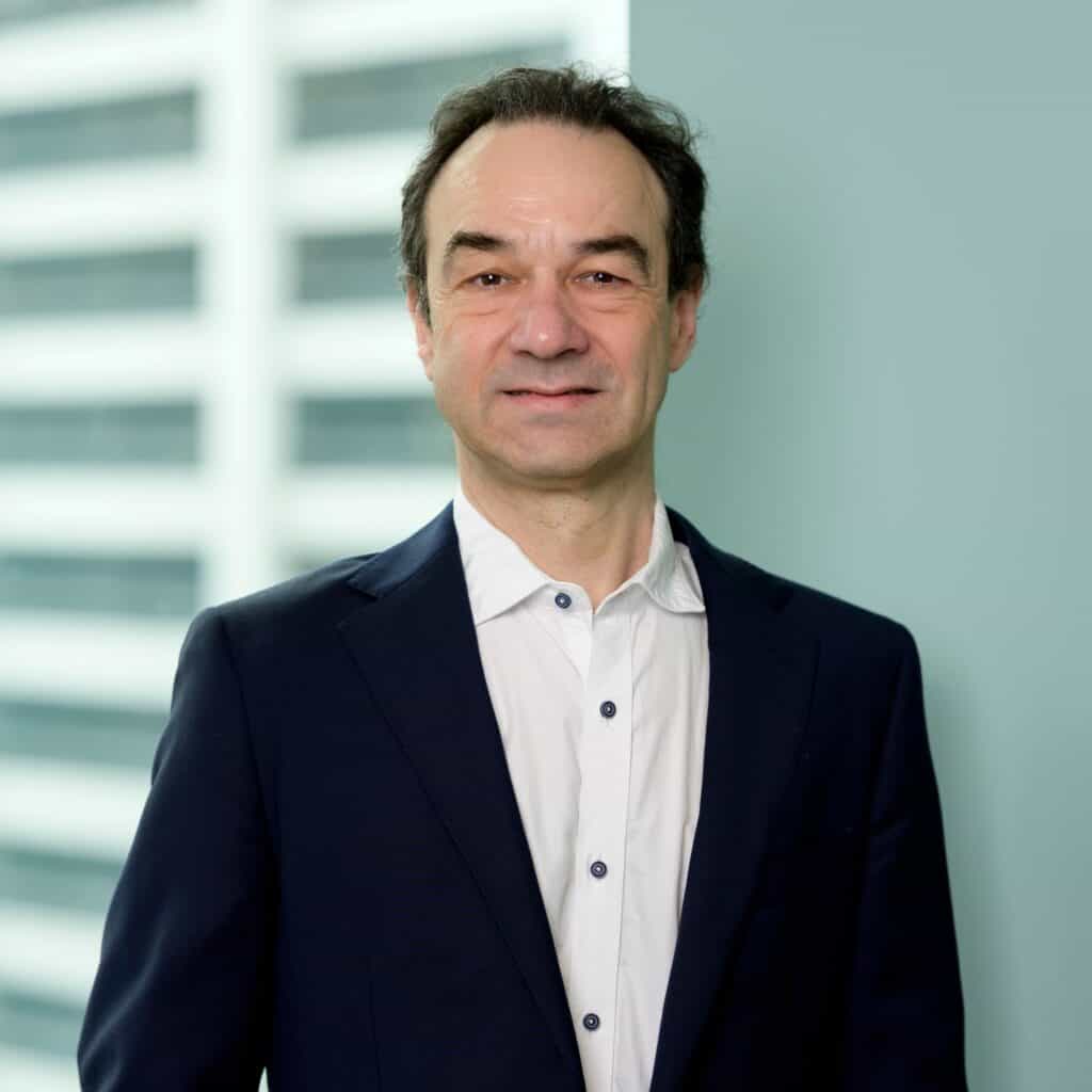 Andrew Tessler, wearing a navy blue suit and white shirt, stands indoors in front of a light blue wall and a blurred window with vertical lines, smiling slightly at the camera.