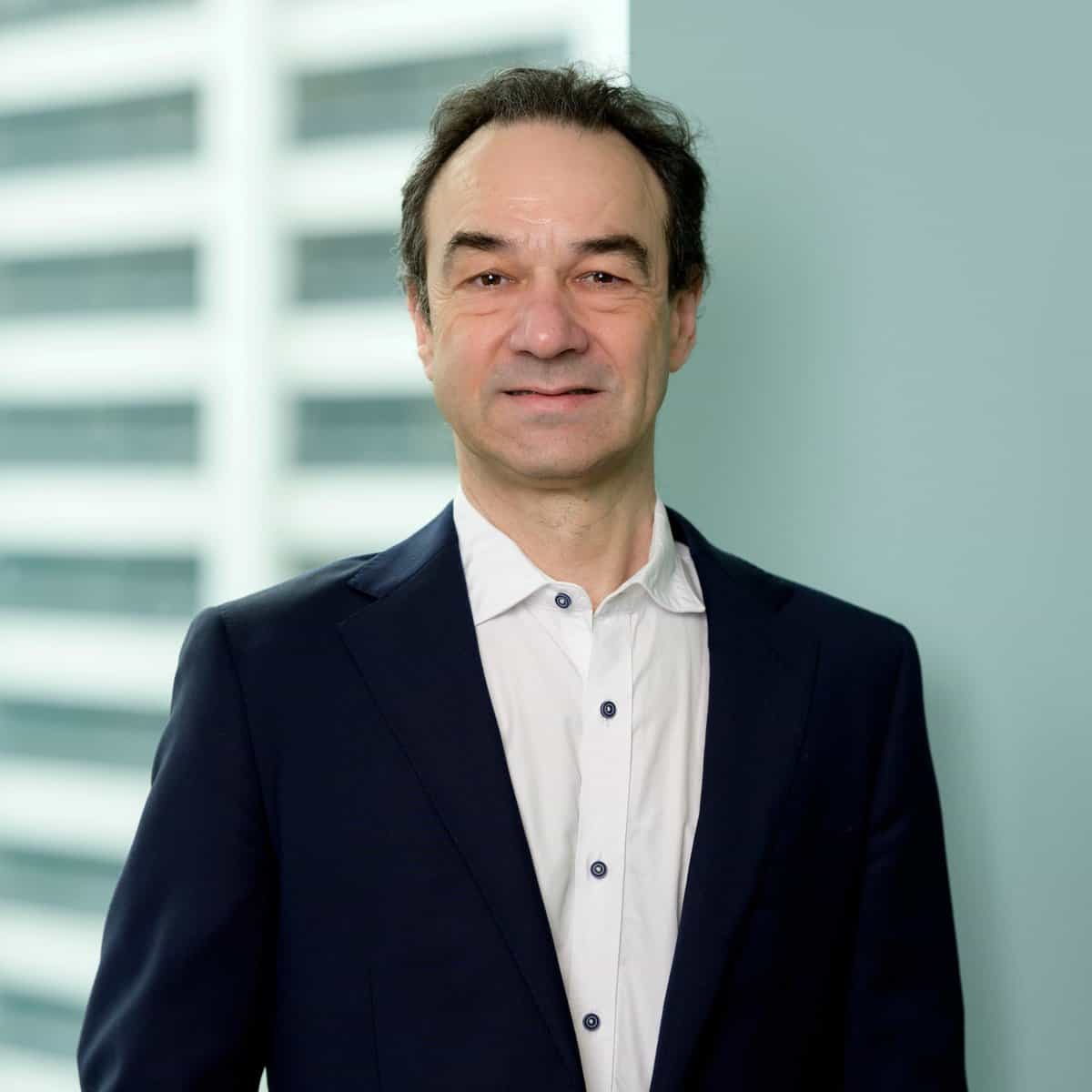 Andrew Tessler, wearing a navy blue suit and white shirt, stands indoors in front of a light blue wall and a blurred window with vertical lines, smiling slightly at the camera.
