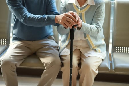 Two elderly people sit side by side on a bench, one offering a comforting hand over the other's hands, which rest on a walking cane—a quiet moment of care that echoes gentle governance and support. Their faces are not visible.