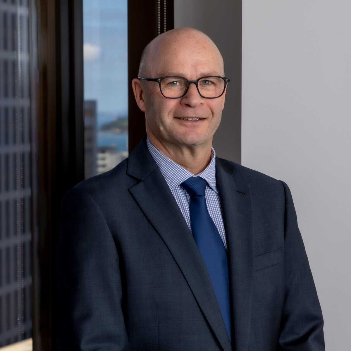 A man wearing glasses, a dark suit, blue tie, and checked shirt stands indoors near a window with city buildings and a blue sky visible in the background.