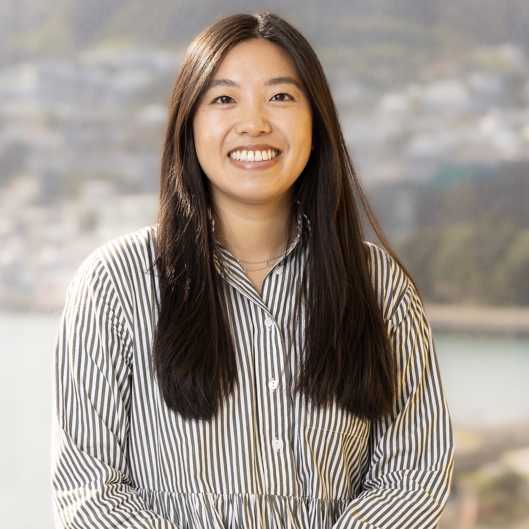 A woman with long dark hair wearing a striped button-up shirt smiles at the camera. The background shows a blurry outdoor scene with water and buildings.