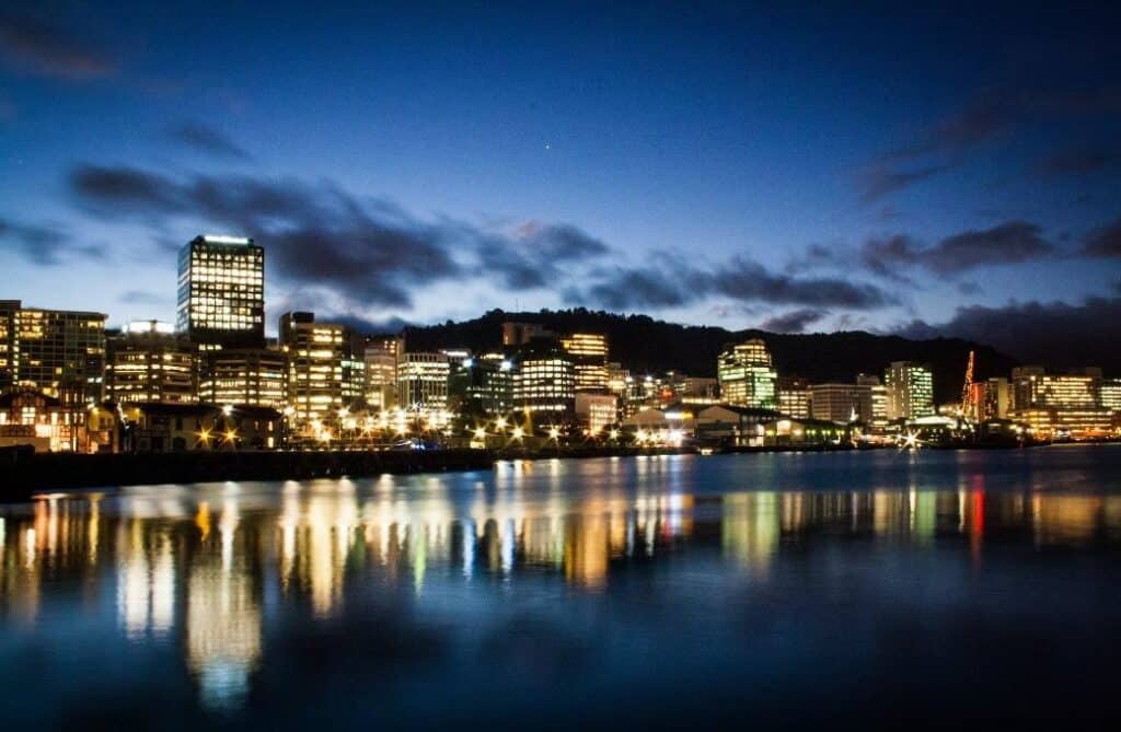 City skyline at dusk with illuminated office buildings reflecting on calm water, solid progress rising above the hills in the background, beneath a dark blue sky with scattered clouds.