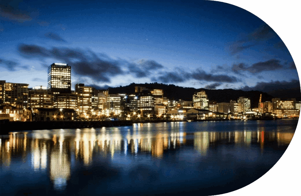 City skyline at dusk with illuminated buildings reflected in calm water, suggesting solid progress against a darkening blue sky with scattered clouds. The image features a curved white border on the top right and lower right edges.