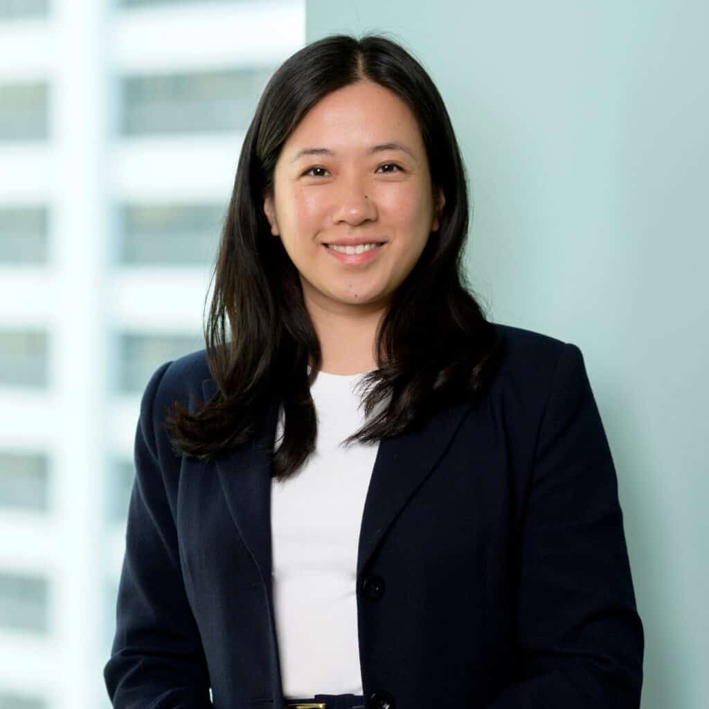 A woman with long dark hair, wearing a navy blazer and white top, smiles while standing indoors against a light blue wall with large windows and city buildings in the background.