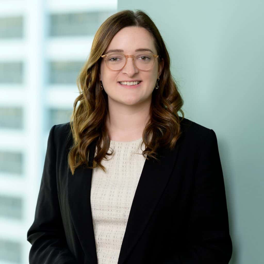 A woman with long brown hair, glasses, and a black blazer over a light-colored top smiles while standing indoors, with blurred windows and a light blue wall in the background.